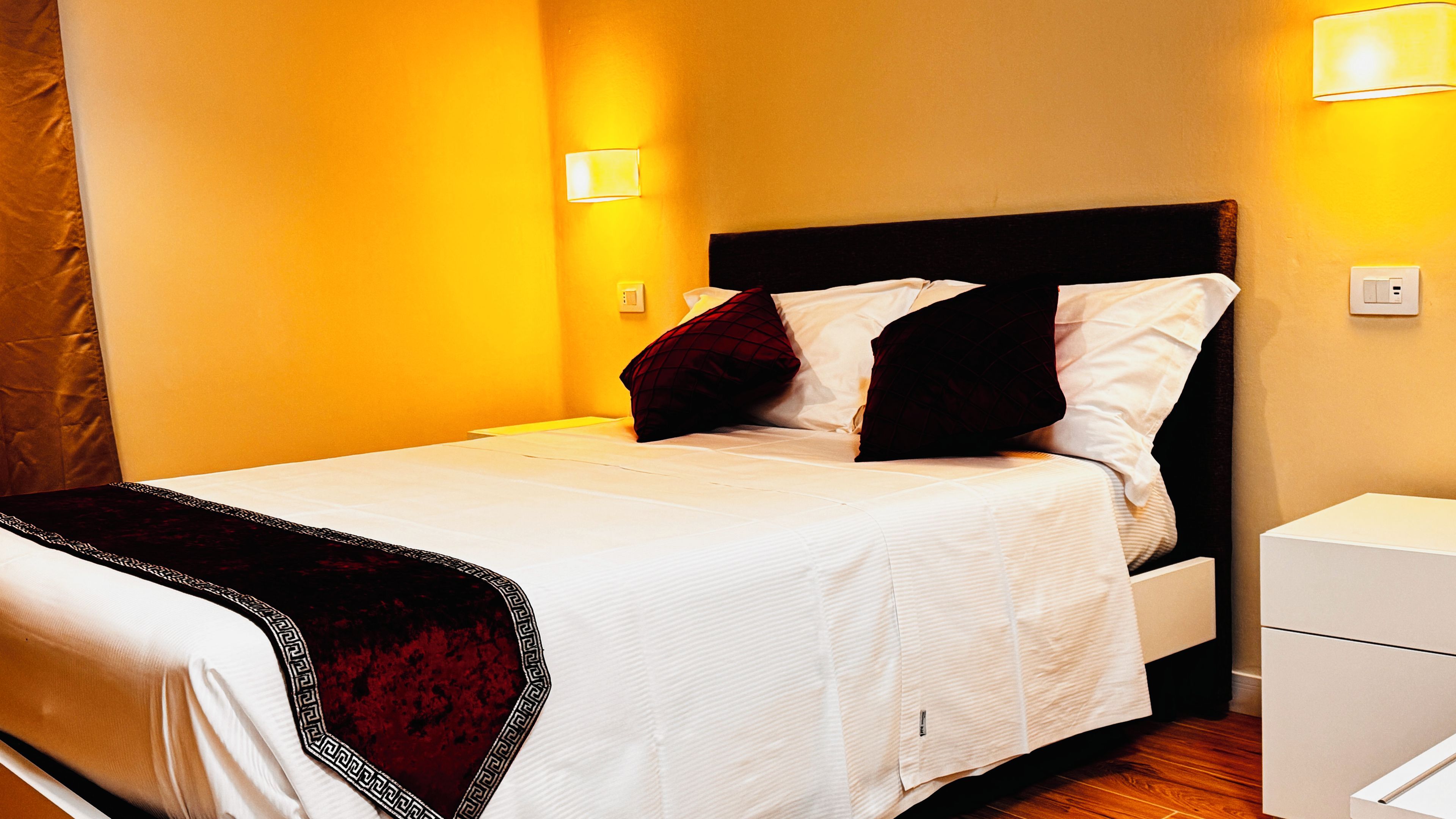 Una cama bien hecha con cabecera oscura, ropa de cama blanca y una manta roja. Hay dos lámparas de noche en la pared.