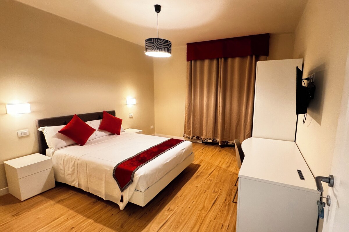 A hotel room with a bed, bedside tables, a TV, and a window with curtains.