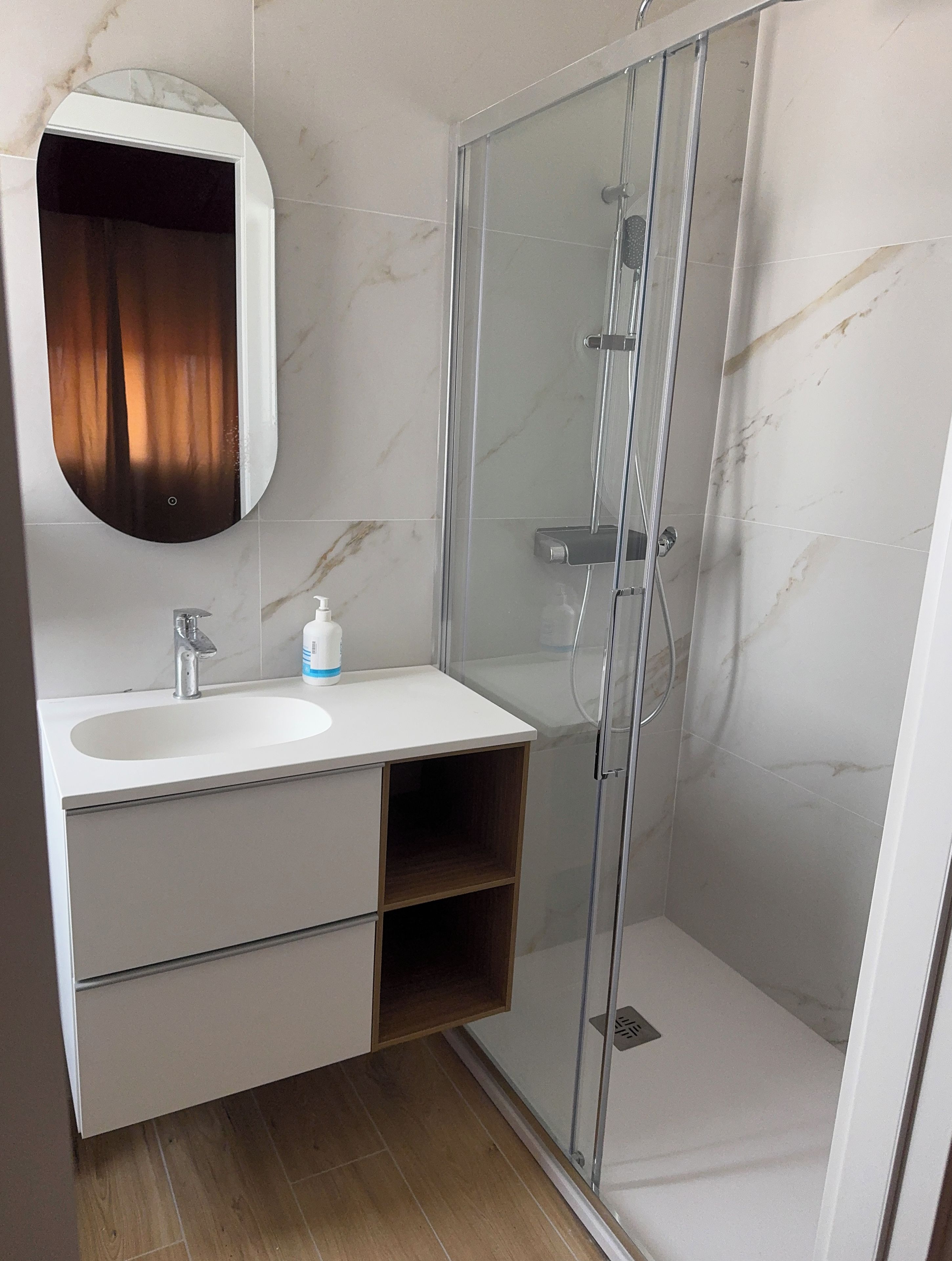 A bathroom with a marble-tiled wall, a white sink with a mirror, a glass-enclosed shower, and wooden storage.