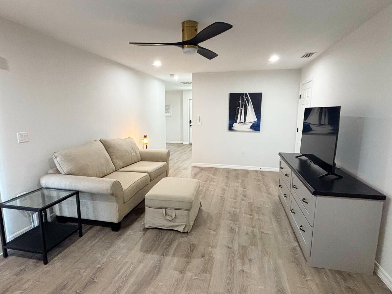 A modern living room with a beige sofa, a glass coffee table, a TV stand with a flat-screen TV, and a ceiling fan.
