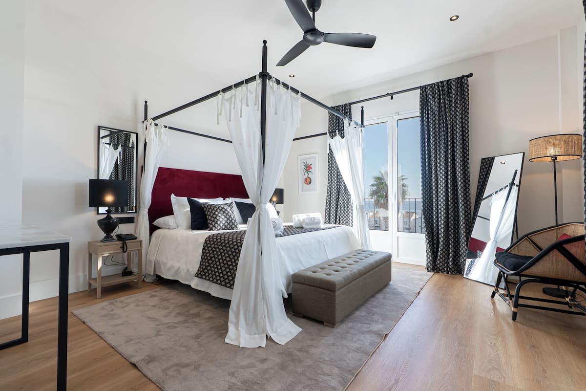 A bedroom with a four-poster bed, a ceiling fan, a bedside table with a lamp, a bench, and a large window with curtains.