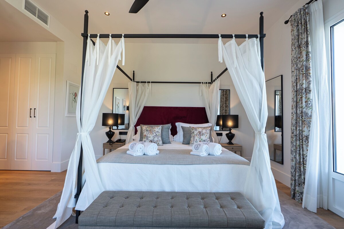 A four-poster canopy bed with a red headboard, bedside tables with lamps, and a grey bench in a well-lit room with large windows.