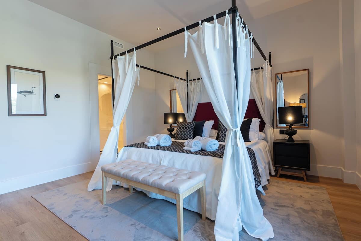 A four-poster canopy bed with rolled towels, a bench, and a bedside table with a lamp.