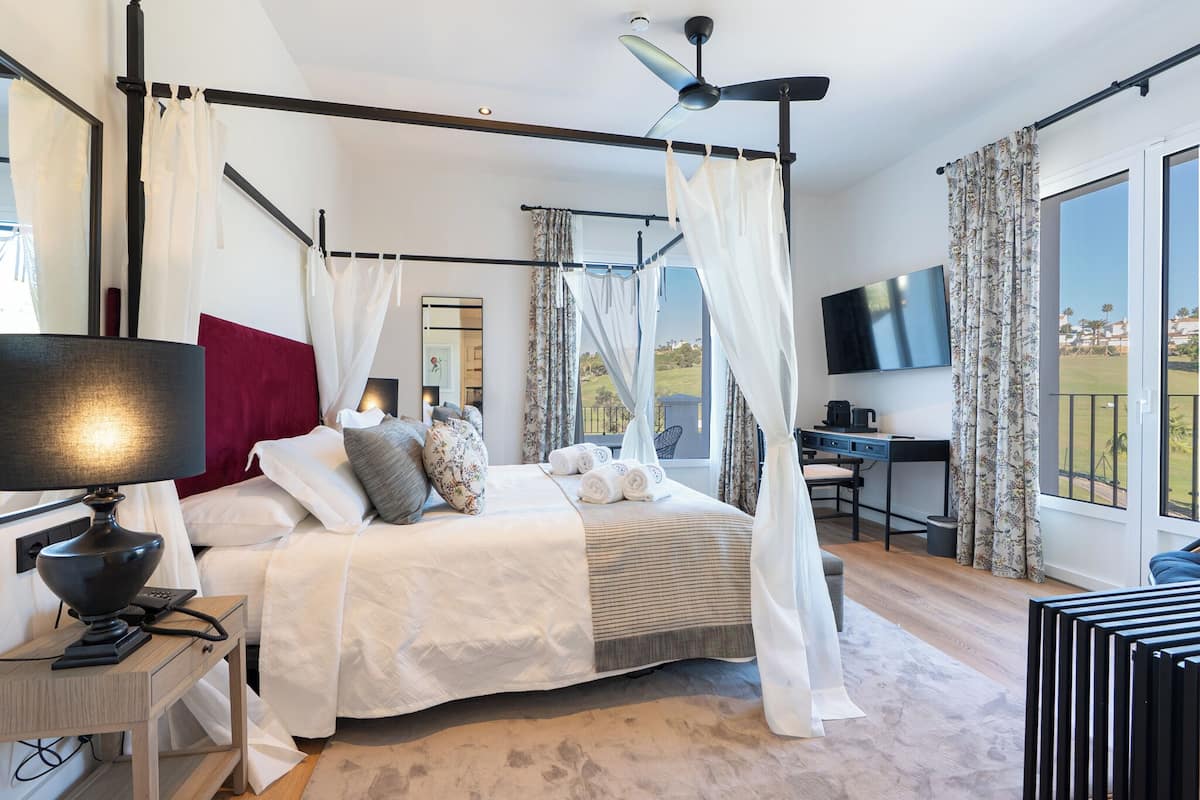 A bedroom with a four-poster bed, a desk, a television, and a view of the outdoors.