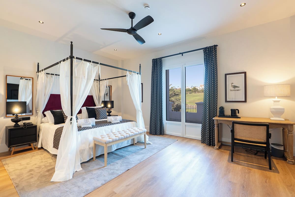 A four-poster bed with a canopy, a desk with a chair, and a view of the outdoors.
