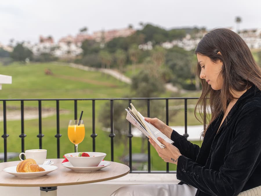 A person sitting at a table with a croissant, coffee, and a glass of orange juice, reading a book.