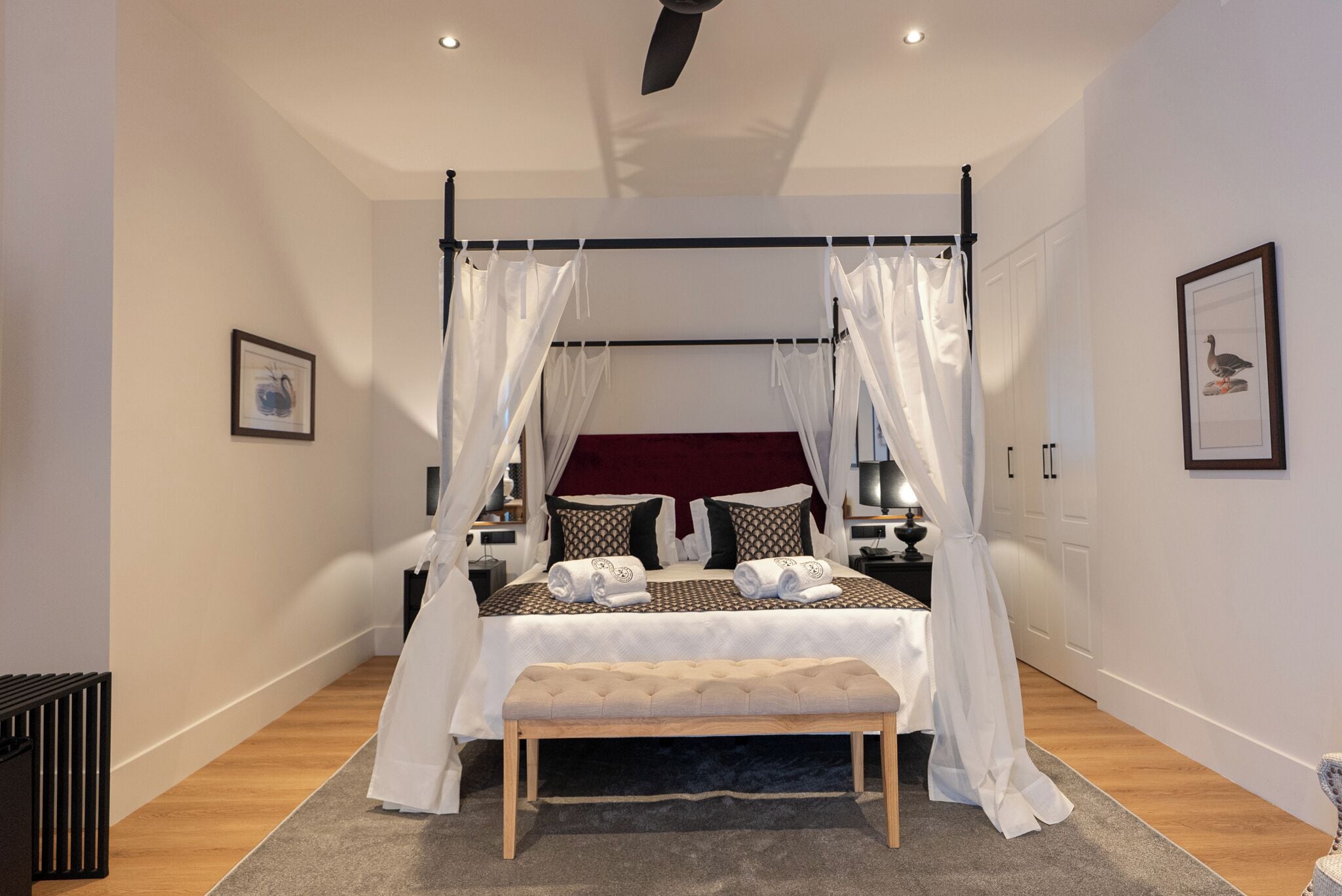 A four-poster bed with white drapes, a bench, and a bedside table with a lamp.
