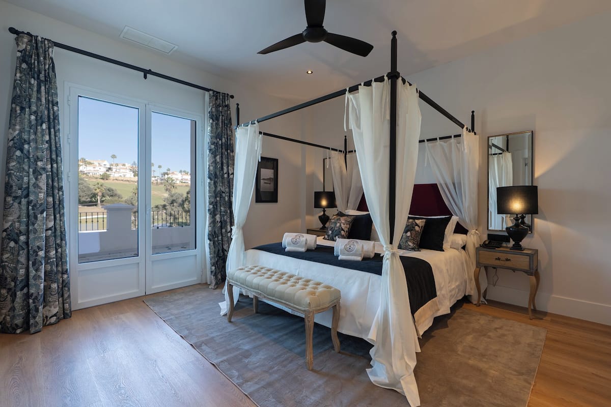 A bedroom with a four-poster bed, a bench, a bedside table with a lamp, and a large window with curtains.