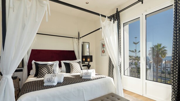 A bedroom with a large bed, a bedside table, a lamp, a window with a view, and a balcony.