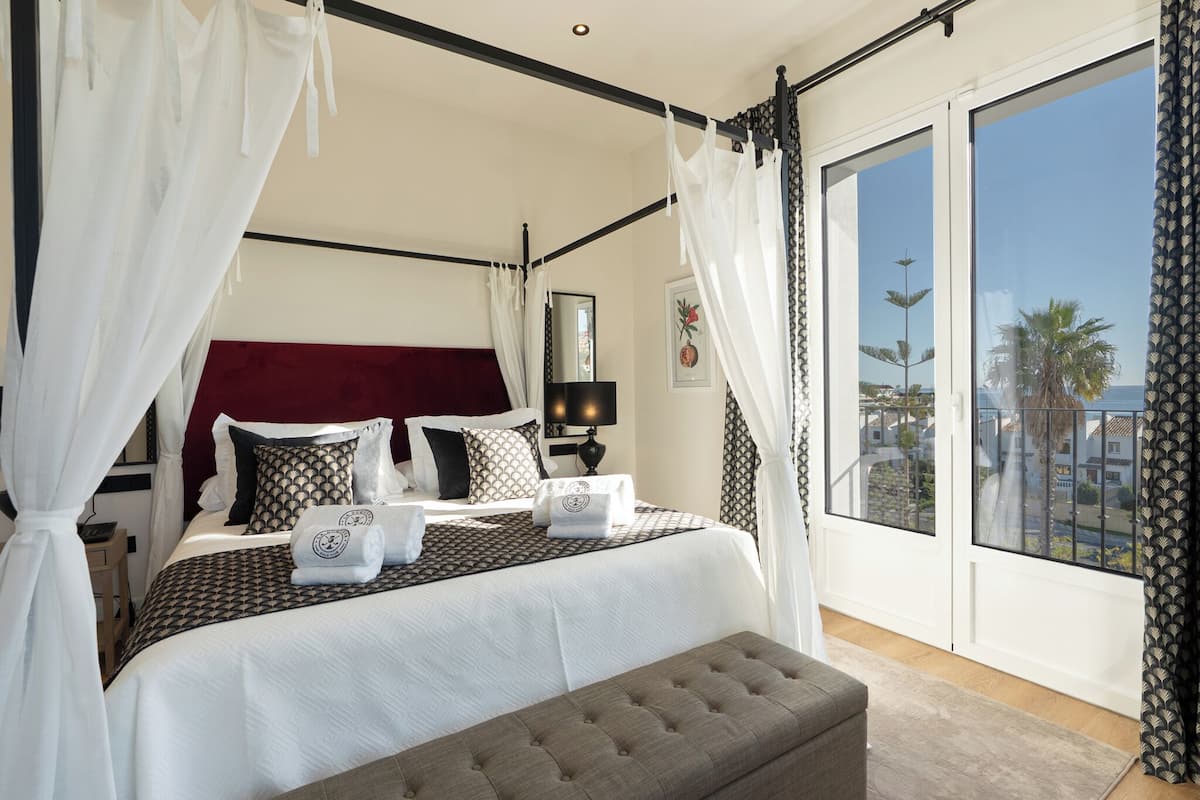 A bedroom with a large bed, a bedside table, a lamp, a window with a view, and a balcony.