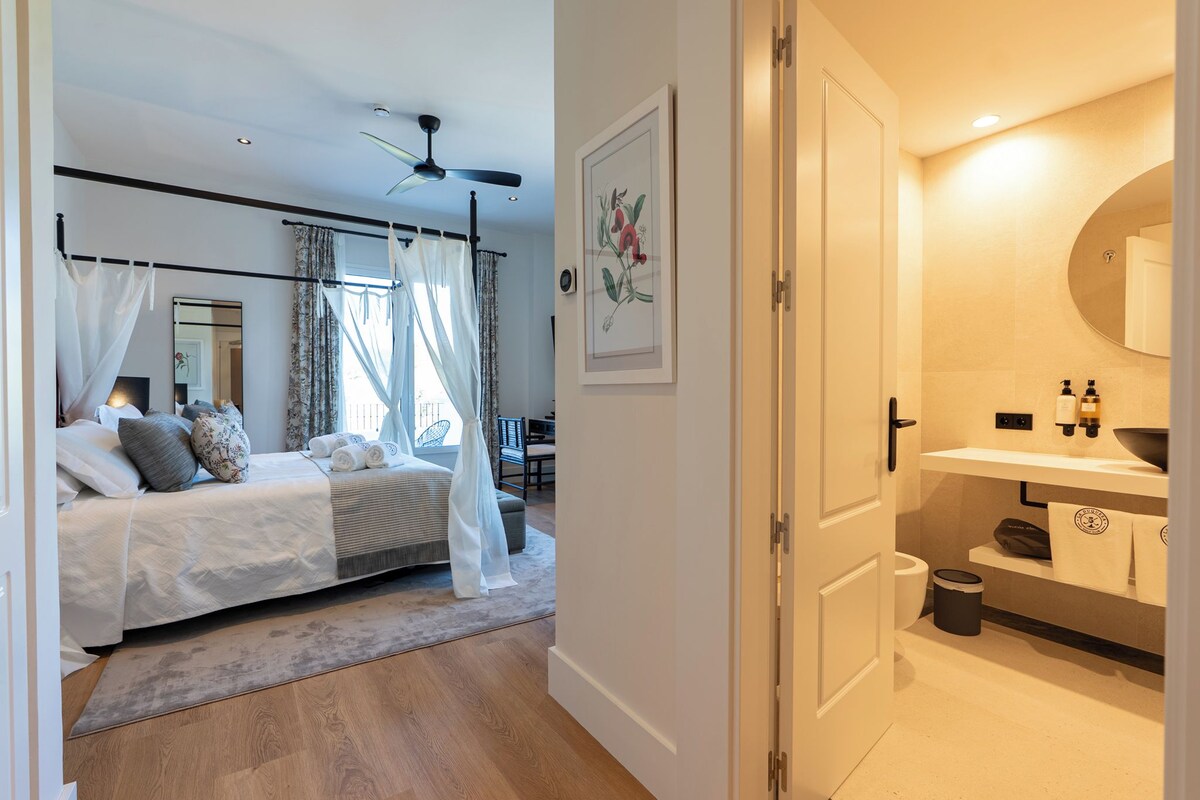 A bedroom with a bed, a ceiling fan, and a bathroom with a sink and mirror.