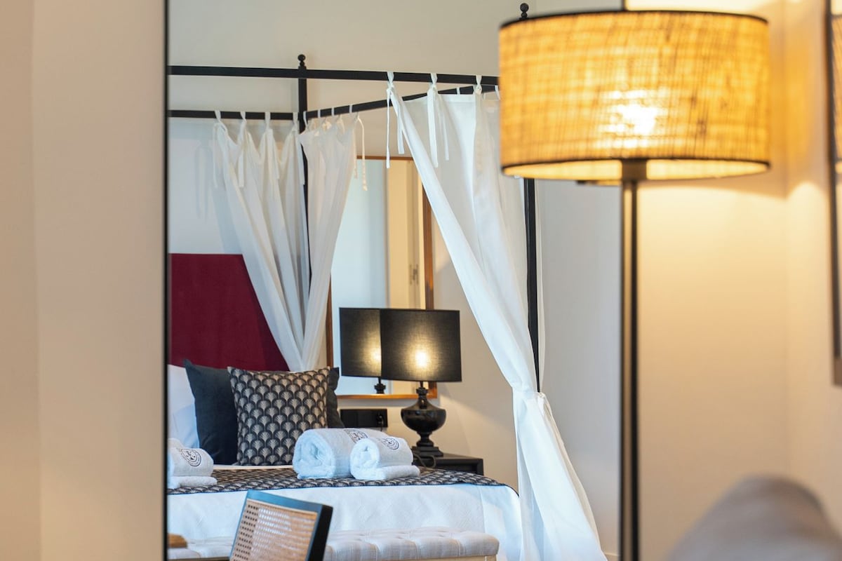 A bedroom with a four-poster bed, a bedside lamp, and a mirror reflecting the room.