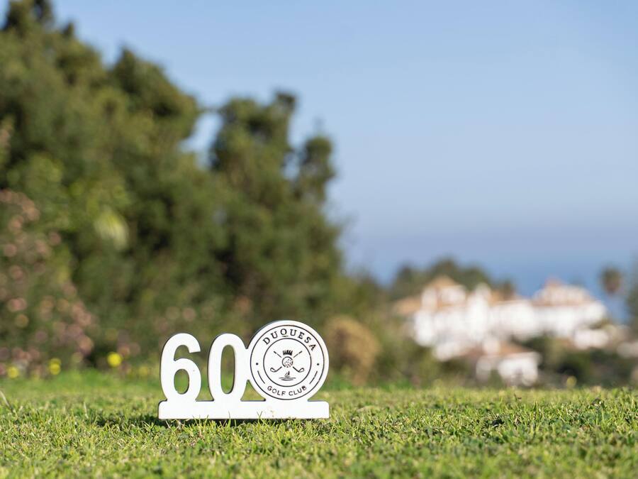Señal de club de golf con el número 60 y un logotipo, ubicada en un área cubierta de césped con árboles y una vista distante de casas.