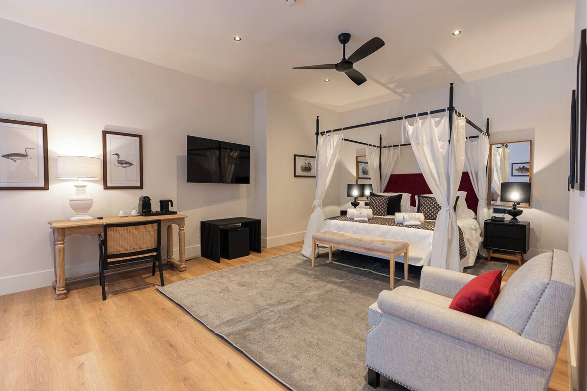 A bedroom with a four-poster bed, a ceiling fan, a TV, a bedside table with a lamp, and a sofa.