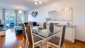 Moderni ruokailutila, jossa on lasipöytä ja valkoiset tuolit, kattokruunu sekä suuri ikkuna, josta on kaunis näkymä.
