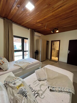 A hotel room with two beds, wooden ceiling, large windows, and an air conditioning unit.