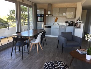 A modern kitchen with white cabinets, a built-in oven, and a coffee machine. There is a dining area with a wooden table and chairs, and a grey sofa.