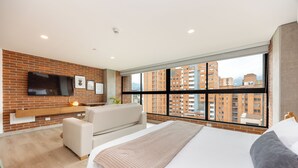 Una habitación de hotel moderna con una cama grande, un sofá, un televisor montado en una pared de ladrillo y un ventanal con vistas a la ciudad.