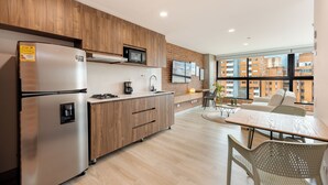 Cocina moderna con armarios de madera, un refrigerador de acero inoxidable, un microondas y una cafetera. Hay un comedor con una mesa y sillas, y un amplio ventanal con vistas a edificios.