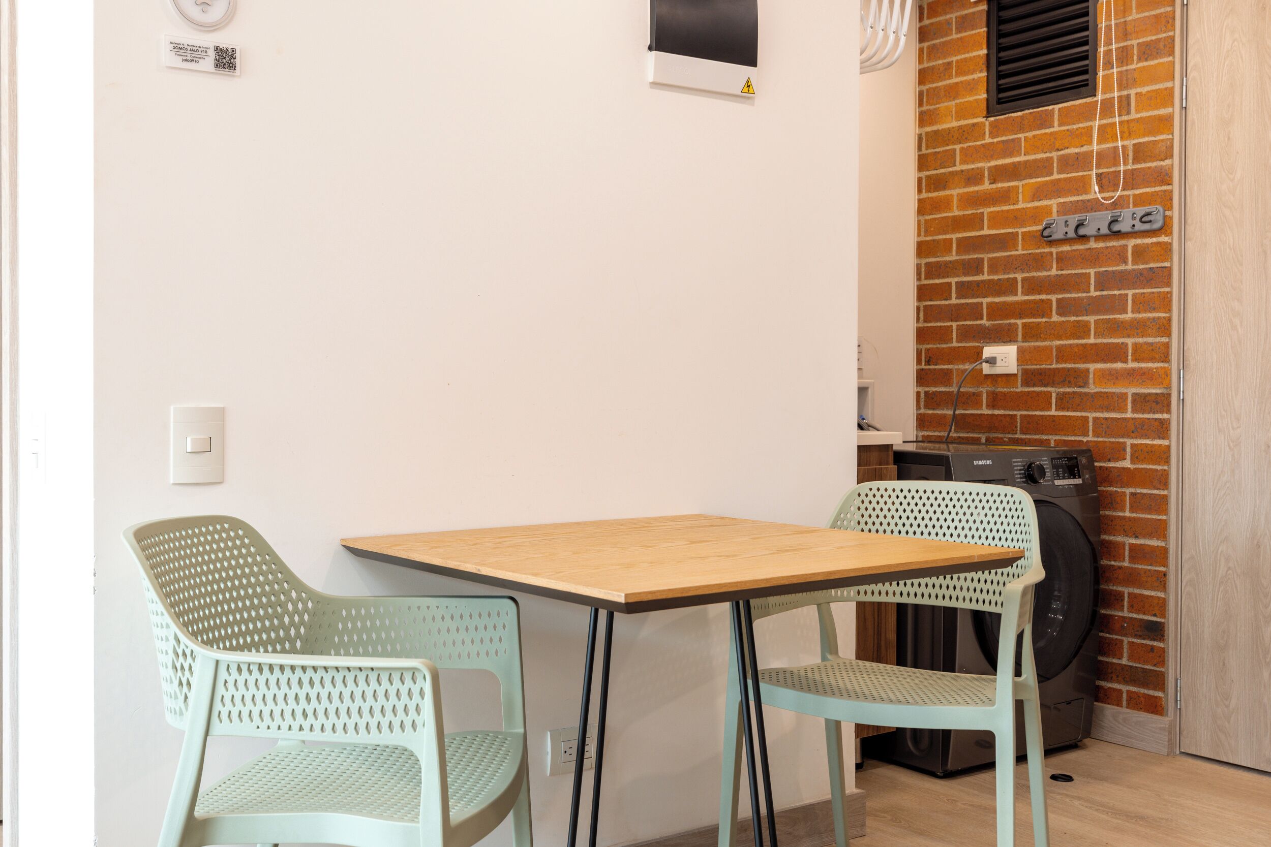 Una habitación pequeña con una mesa de madera y dos sillas verdes. Se observa una lavadora y una pared de ladrillo al fondo.