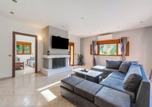 Un salón espacioso con un sofá grande de color gris, un televisor de pantalla plana montado sobre una chimenea, y un comedor visible a través de un umbral abierto.
