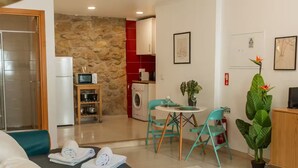 Un espacio habitable compacto con kitchenette, una zona de comedor con mesa y sillas, un sofá y una planta grande en maceta.