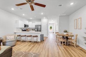 A modern kitchen with white cabinets, a central island, and a dining area with wooden chairs. - New Construction: Coastal Retreat • 5 Mins to Downtown OS (Ocean Springs)