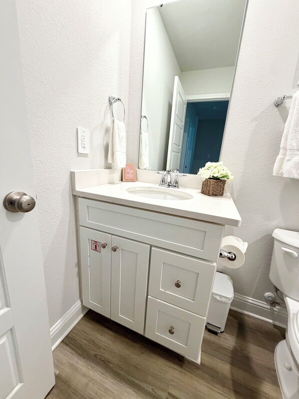 A bathroom with a white sink, mirror, and toilet. There are towels hanging on the wall and a small plant on the counter. - New Construction: Coastal Retreat • 5 Mins to Downtown OS (Ocean Springs)
