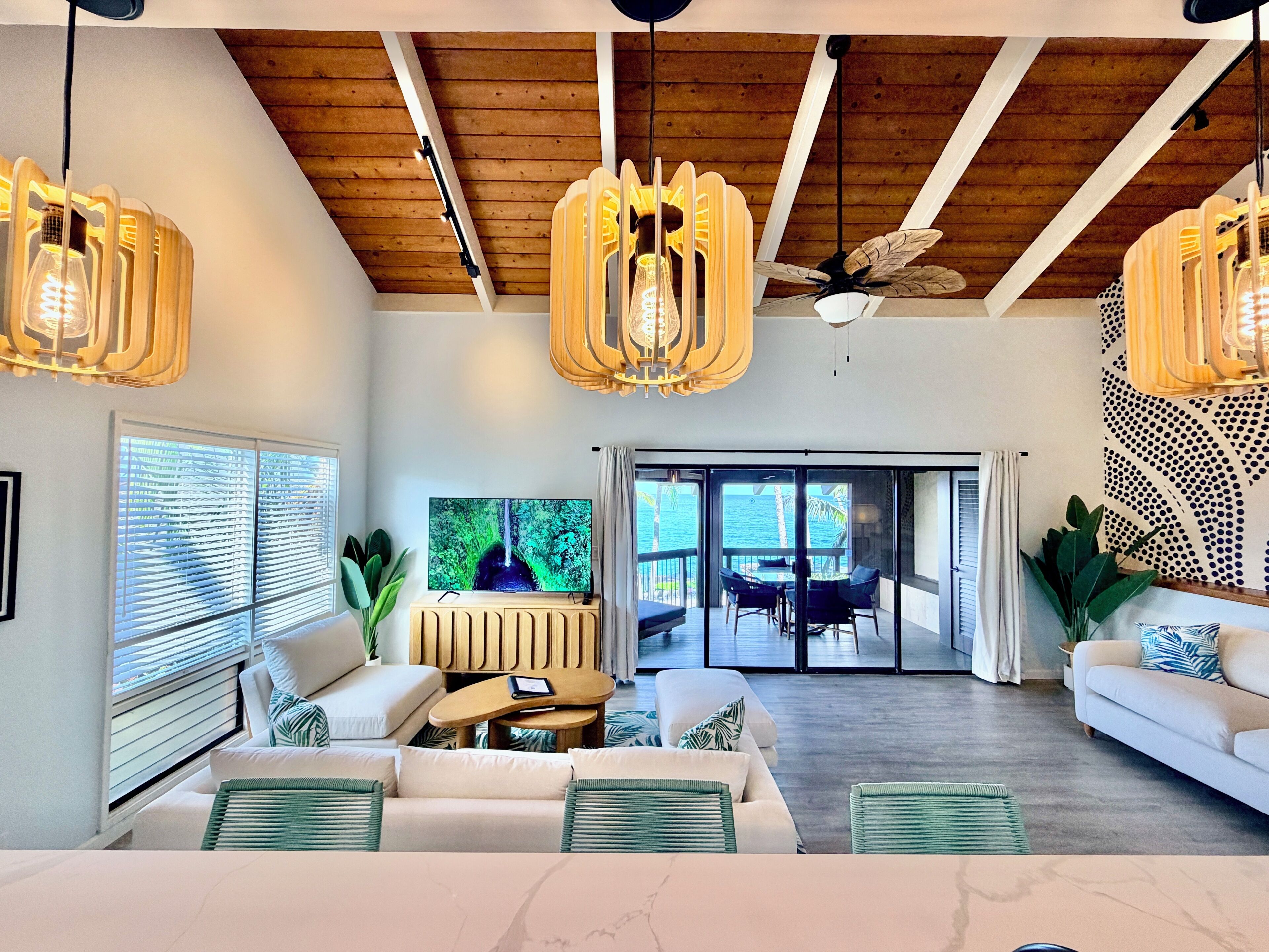 A modern living room with a large TV, wooden ceiling beams, and a view to the outside.