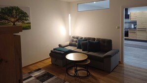 Un salon moderne avec un canapé, un pouf et une table basse ronde. Un tableau représentant un arbre est accroché au mur, et une fenêtre laisse entrer la lumière naturelle.