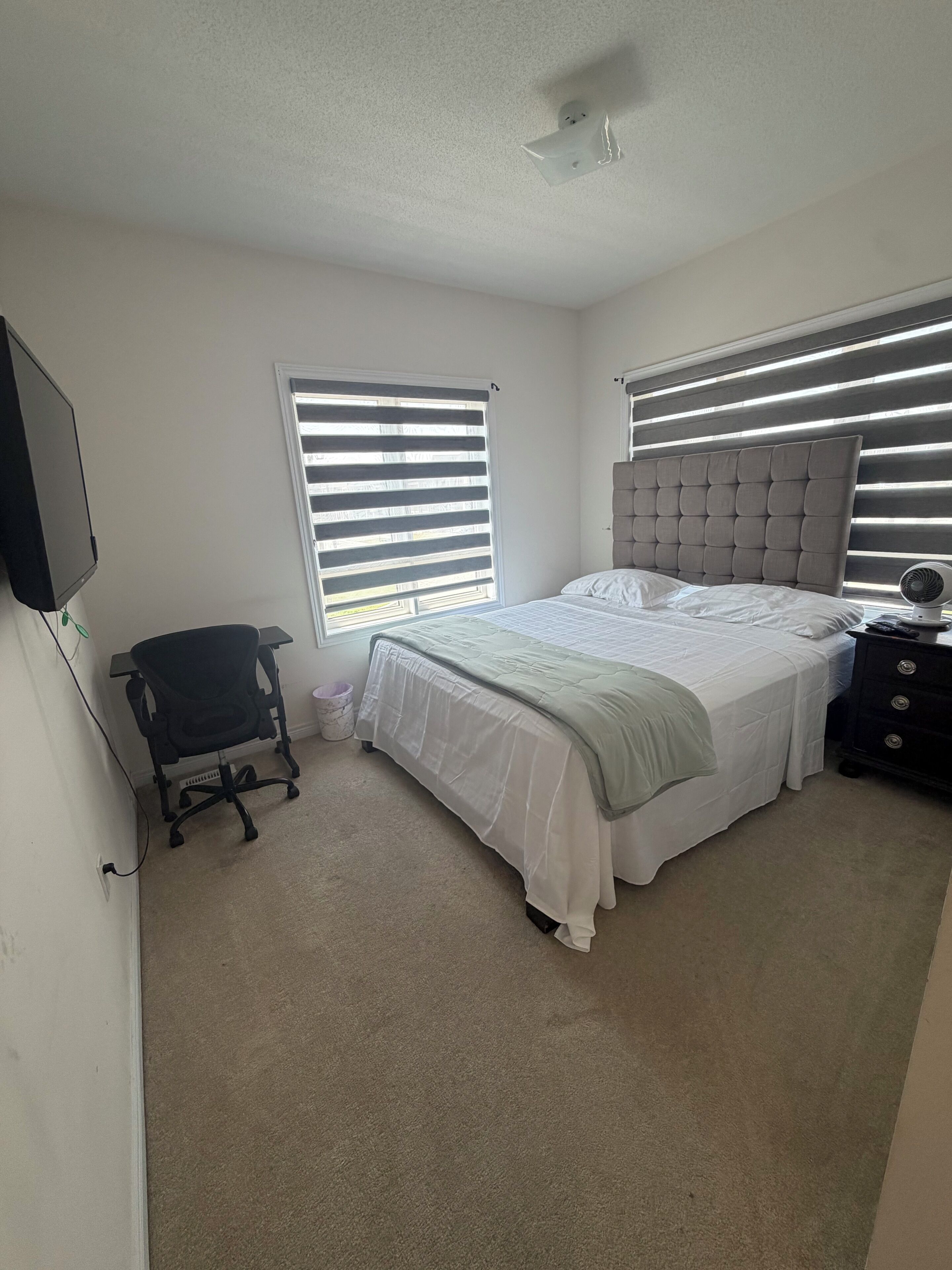 A bedroom with a bed, a chair, a bedside table, a television, and a window with blinds.