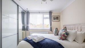 Een slaapkamer met een bed, een blauwe deken, witte kussens, een kleurrijk kussen, een raam met gordijnen en een foto aan de muur.