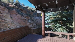 Ett trädäck med en pergola ovanför, en mur av tegelsten och utsikt över en bergssluttning med sparsam växtlighet.