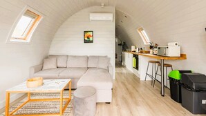 Een compacte, loft-stijl leefruimte met een bank, een klein tafeltje en een keukenhoek met krukjes.