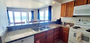 A kitchen with granite countertops, a stainless steel sink, wooden cabinets, a white stove, and a view of the ocean through large windows. - Condo #208 with beautiful sunset view from your balcony overlooking the ocean (Lincoln City)