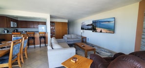 A modern living room with a sofa, armchair, and coffee table, a kitchen with wooden cabinets and a refrigerator, and a large painting of a beach scene on the wall. - Condo #208 with beautiful sunset view from your balcony overlooking the ocean (Lincoln City)