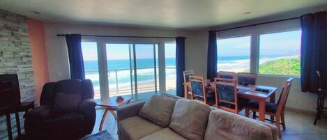 A living room with a view of the beach, a sofa, a TV, and a dining area with chairs.
