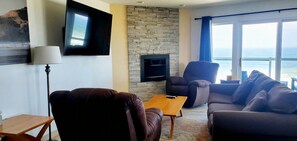 A living room with a stone wall, a flat-screen TV, leather sofas, and a view of the sea. - Condo #308 with beautiful sunset view from your balcony overlooking the ocean (Lincoln City)