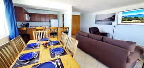 A dining area with a wooden table set for four, a brown sofa, and a kitchen in the background. - Condo #308 with beautiful sunset view from your balcony overlooking the ocean (Lincoln City)