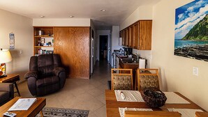 Ein Wohnzimmer mit Ledersessel, Couchtisch mit Zeitung, Bücherregal und Küchenzeile mit Holzschränken sowie einem Gemälde von einer Küstenlandschaft.