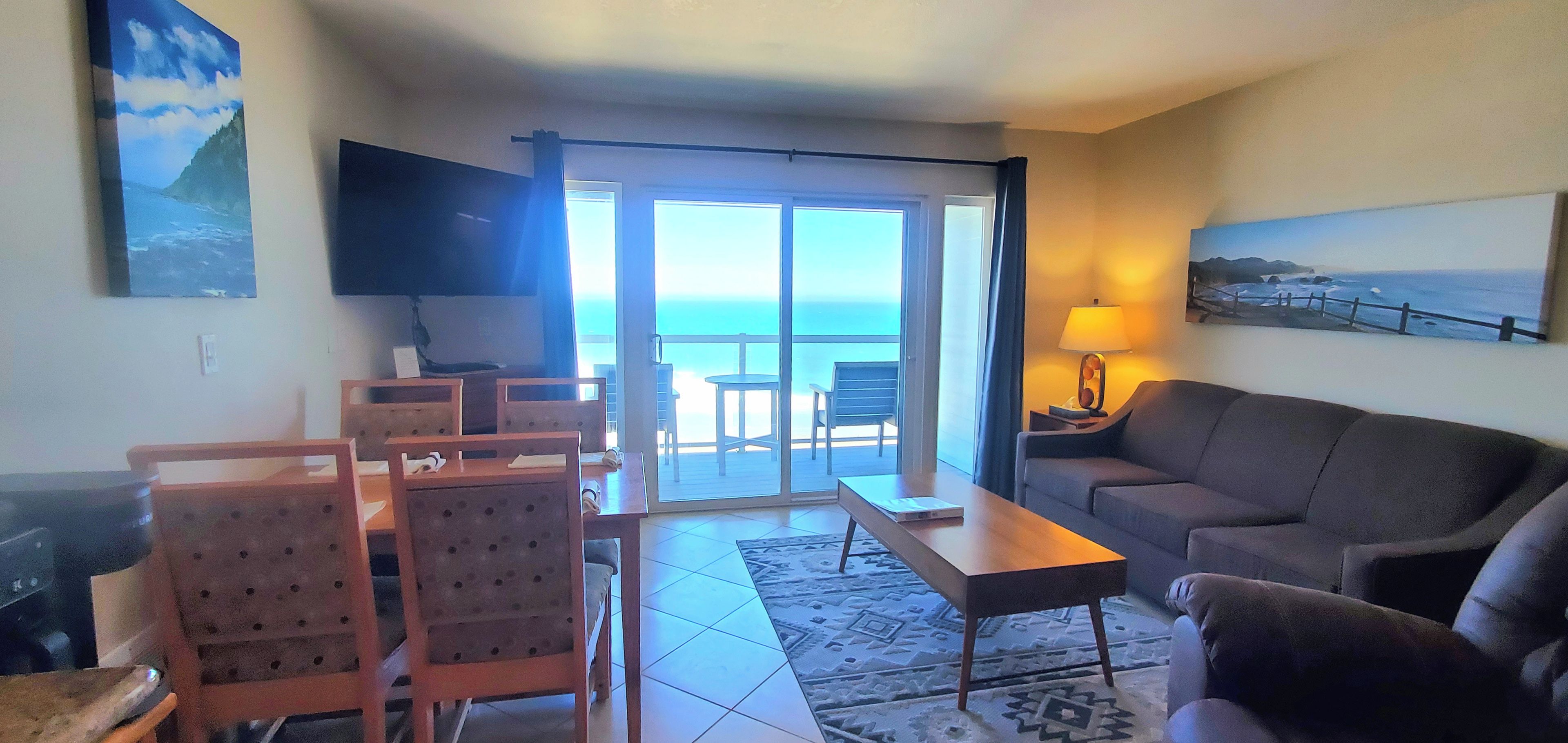 A living room with a sofa, a TV, a dining table with chairs, and a view of the ocean through a sliding door.
