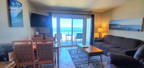 A living room with a sofa, a TV, a dining table with chairs, and a view of the ocean through a sliding door. - Condo #202 with beautiful sunset view from your balcony overlooking the ocean (Lincoln City)