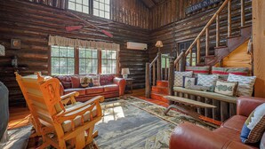 A rustic cabin interior with a wooden staircase, a leather sofa, a rocking chair, and a large window. - Quintessential Maine Lake House, 280Ft of Waterfront, near Acadia & Bar Harbor (Dedham)