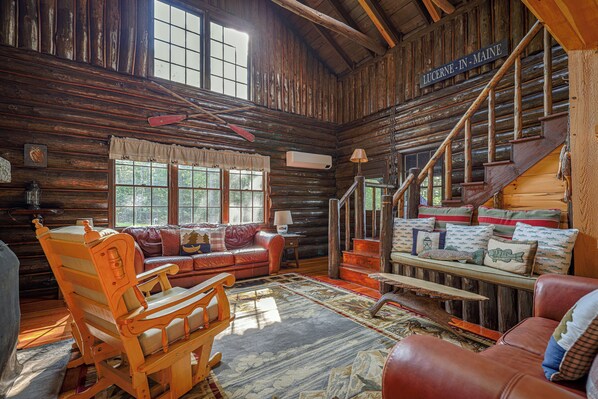 A rustic cabin interior with a wooden staircase, a leather sofa, a rocking chair, and a large window.