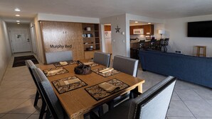 Een woonkamer met een Murphy bed, een eethoek met een houten tafel en stoelen, en een keuken op de achtergrond.