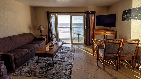 A living room with a sofa, coffee table, and chairs, a TV, and a view of the ocean through a sliding door. - Condo #204 with beautiful sunset view from your balcony overlooking the ocean (Lincoln City)