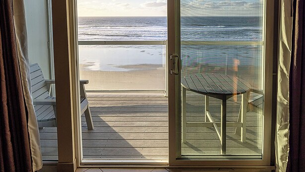 A room with a view of the beach, a sliding glass door, a small table, and a bench. - Condo #204 with beautiful sunset view from your balcony overlooking the ocean (Lincoln City)