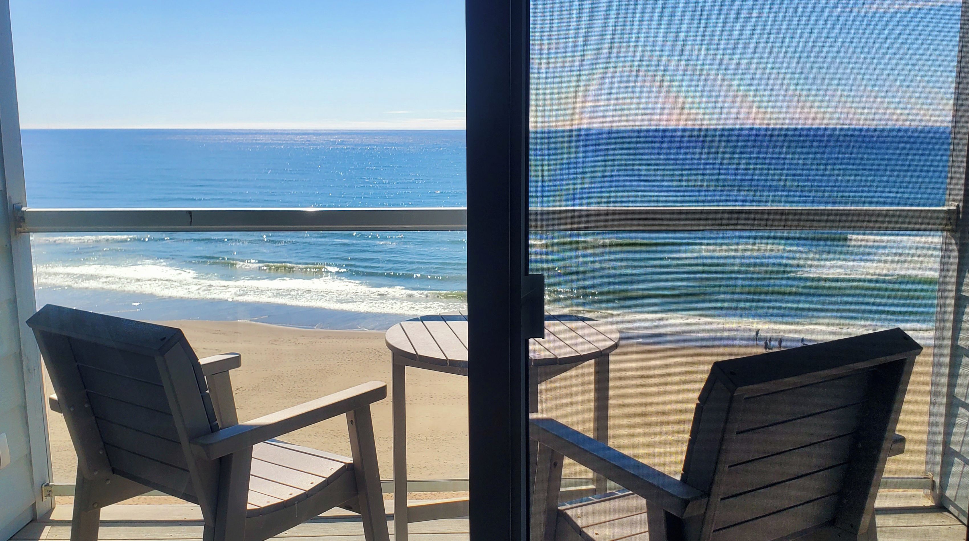 A balcony with two chairs and a small table, overlooking a beach with waves and a clear sky.