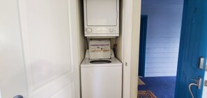 A compact, white laundry room with a built-in washer and dryer, a door leading to a hallway, and a blue door visible in the background. - Condo #108 with beautiful sunset view from your balcony overlooking the ocean (Lincoln City)