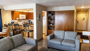 Ein Wohnzimmer mit zwei grauen Sofas, einem Couchtisch und einem Essbereich mit Holzsesseln, sowie eine Küche mit Holzausbau und einer weißen Mikrowelle.